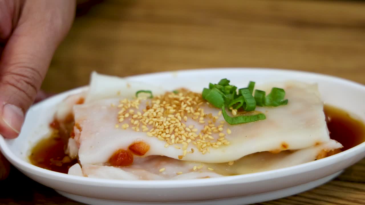 A hand places a rice noodle roll topped with sesame seeds and green onions onto a wooden table, under soft, natural lighting with a steady camera angle