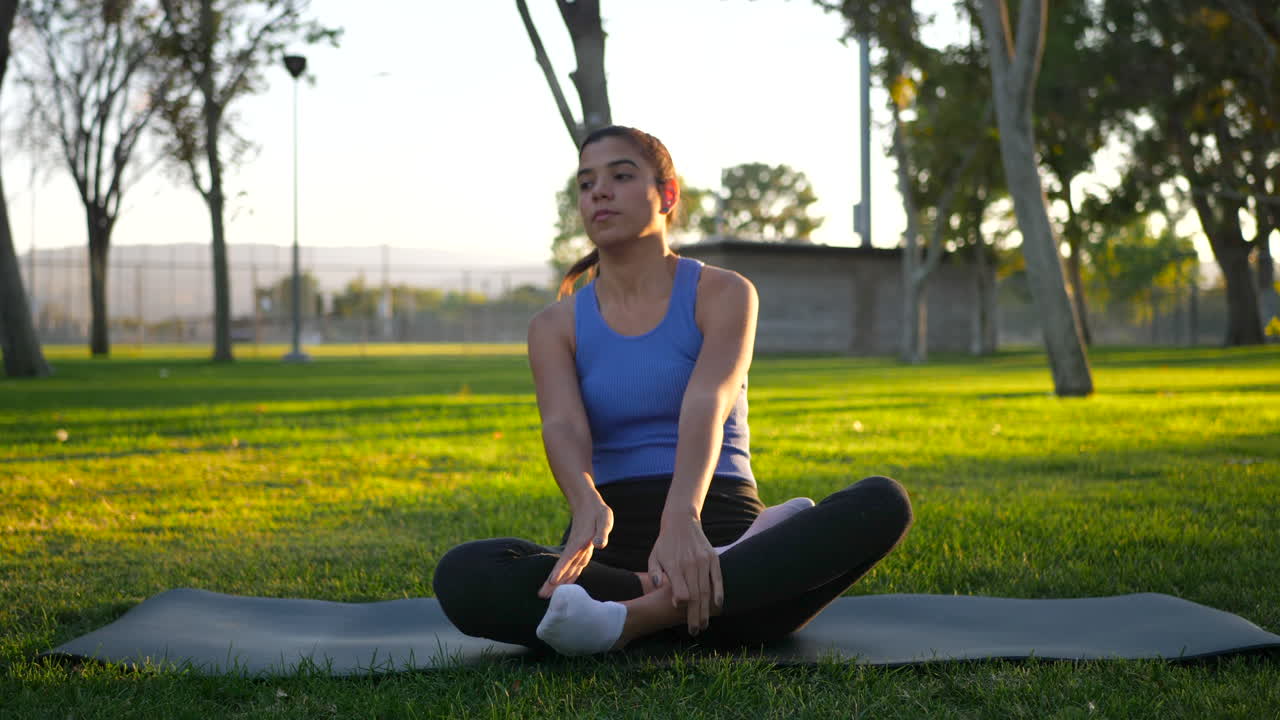 hermosa joven sentada en posición de loto en su alfombra de yoga en meditación durante el amanecer en un parque de césped