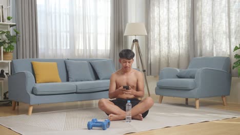 집에서 셔츠를 벗고 운동한 후 덤벨과 함께 매트에 앉아 스마트폰을 사용하는 아시아 십대 소년의 몸이 가득 차 있습니다.