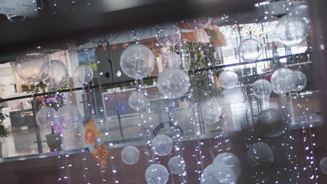 close-up of festive decorations featuring translucent spheres suspended with sparkling string lights, blurred reflections of people moving across glass surfaces, and vibrant greenery