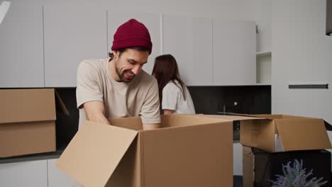 A-happy-man-with-stubble-in-a-red-cap-and-beige-T-shirt-gives-his-brunette-girlfriend-in-a-white-T-shirt-things-from-a-box-after-moving-into-a-new-apartment.-Happy-couple-unpacking-things-from-boxes-after-moving