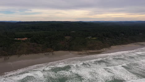 Costa-De-Oregon-Cerca-De-Bandon---Olas-Salpicando-En-La-Playa-De-Arena-Blanca-Con-árboles-Verdes-Durante-La-Puesta-De-Sol---Toma-Aérea