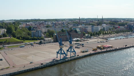 Luftaufnahme-Von-Docks-Mit-Zwei-Hafenkränen.-Glitzernde-Wasseroberfläche-Des-Flusses.-Stadt-Im-Hintergrund