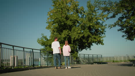 back view of boy in white top and jeans strolling with sister along iron railing on paved path surrounded by trees, benches, and urban structures