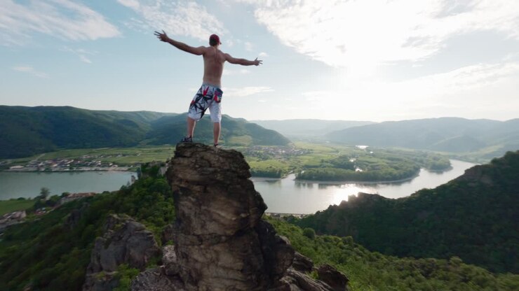 Scalatore in piedi su una roccia, braccia aperte verso il cielo. Movimento del drone FPV attraverso il crinale montuoso e il burrone, posa della vittoria. Bouldering in cima alla vetta, successo spettacolare, ripresa aerea vittoriosa.