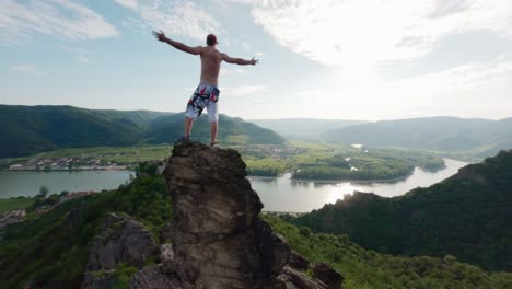 climber-standing-on-rock,-arms-wide-high-in-the-sky,-FPV-drone-movement-through-mountain-ridge-edge-ravine,-victory-pose,-climbing-boulder-on-top-mountaintop-spectaculair-succes-victorious-aerial-shot