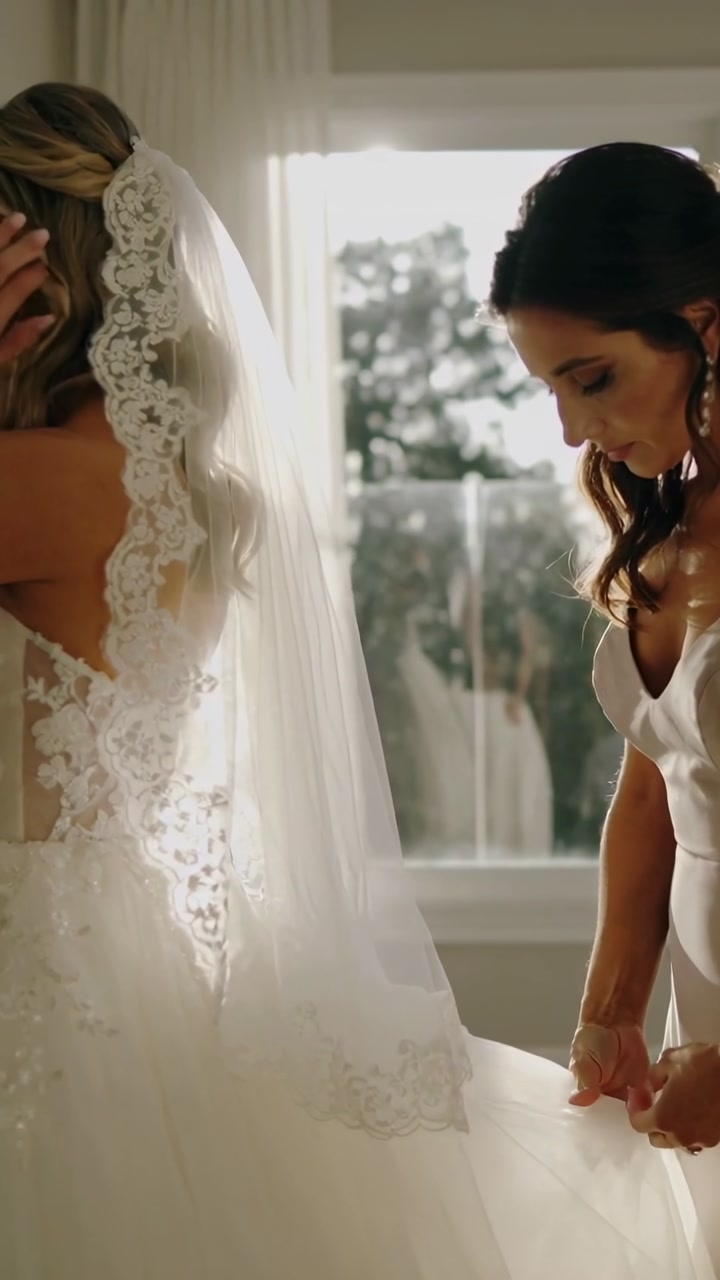 Vertical video: Leaning bridesmaid in satin smoothing lace gown and veil by window, tidying train