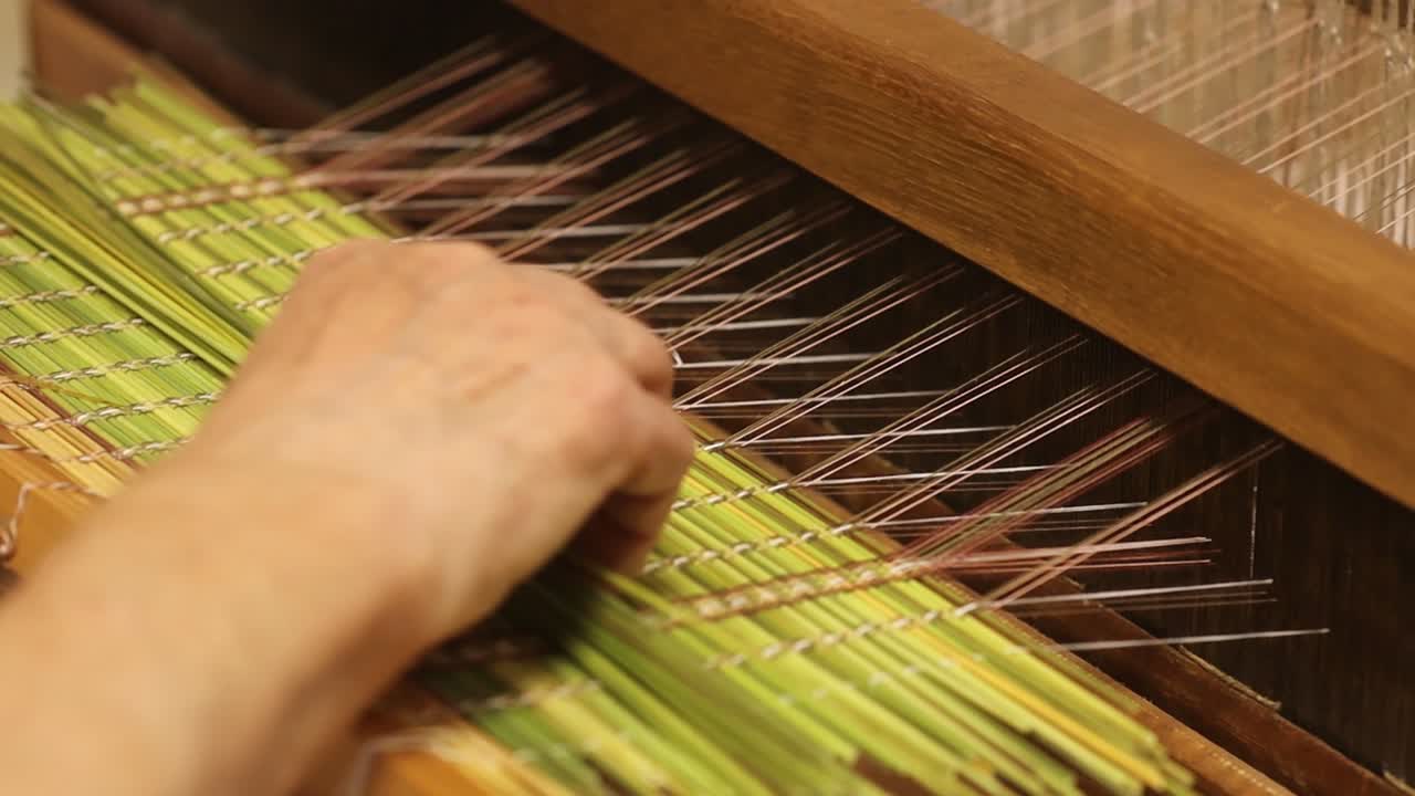 Detailed view of a weaving process using green plant stalks and thin threads on a wooden loom. A hand gently guides the material through the pattern