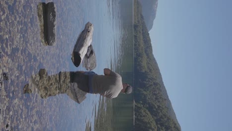 a man with a baseball cap relaxing standing in a lake with clear waters with forests and hills in the backgound