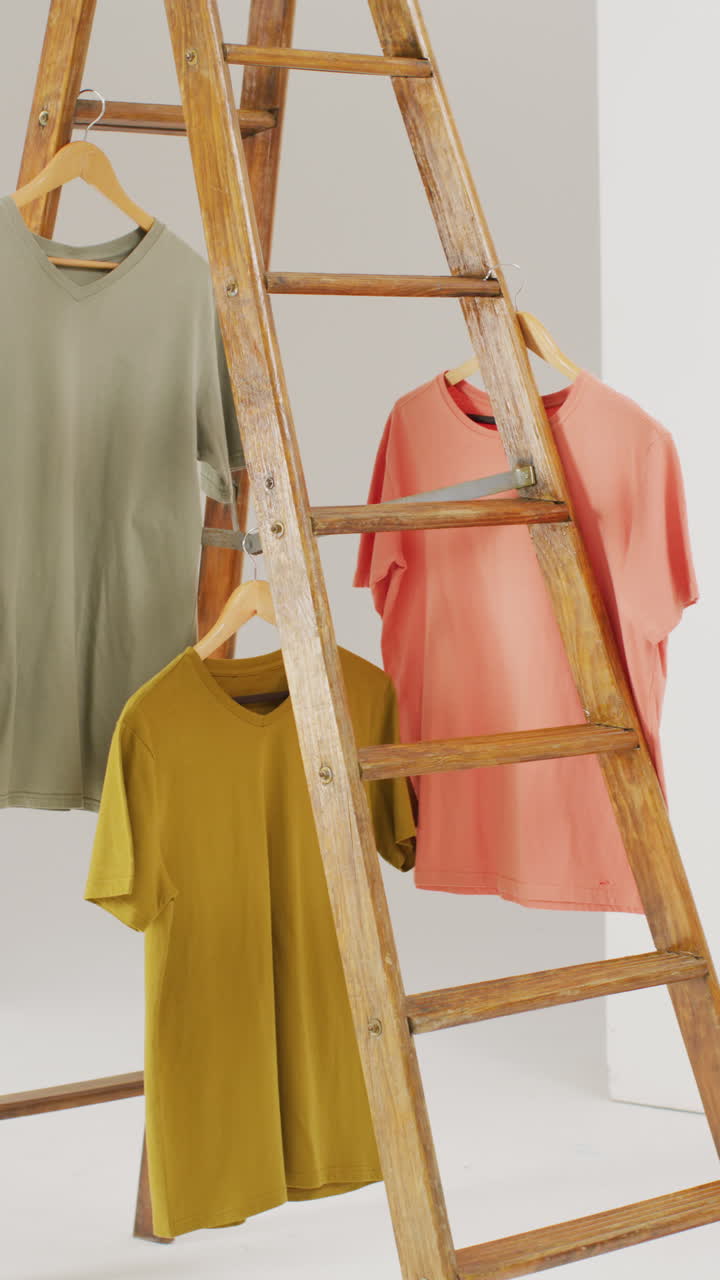 Vertical video of close up of beige, mustard and orange t shirts hanging from ladder with copy space