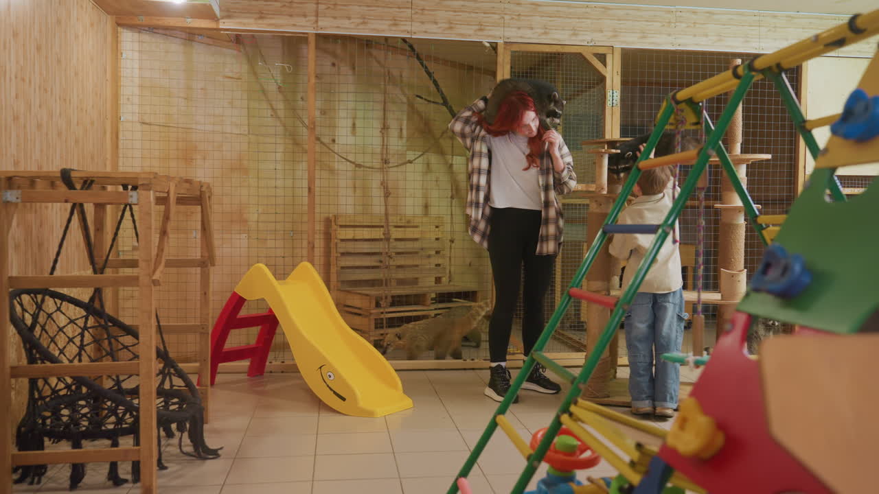 Uma mulher caucasiana ruiva e um menino exploram uma sala de jogos de madeira com tobogãs, estruturas de escalada e torre para gatos, mostrando uma curiosidade divertida enquanto o menino tenta alcançar um gato malhado relaxado, e a mulher ri e ajusta a escalada