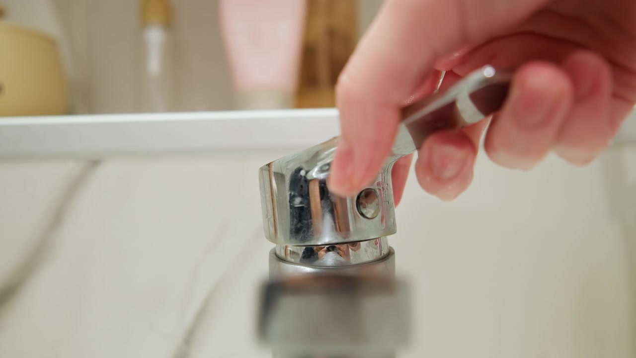 Una vista dal basso mostra una maniglia del rubinetto cromata sollevata quasi orizzontalmente accanto a un lavandino da bagno in marmo, con goccioline d'acqua, metallo riflettente, impugnatura zigrinata e dettagli della cerniera che catturano una luce soffusa e diffusa.