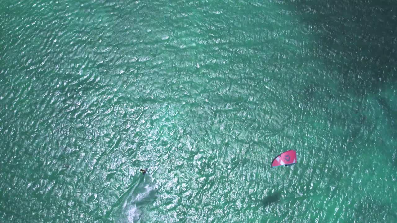 Kitesurfer gliding over clear turquoise water with a red kite, aerial view