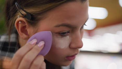 close-up view of young lady applying powder under her eyes with purple makeup sponge during daily beauty routine, focusing on smooth and precise application with professional makeup tools