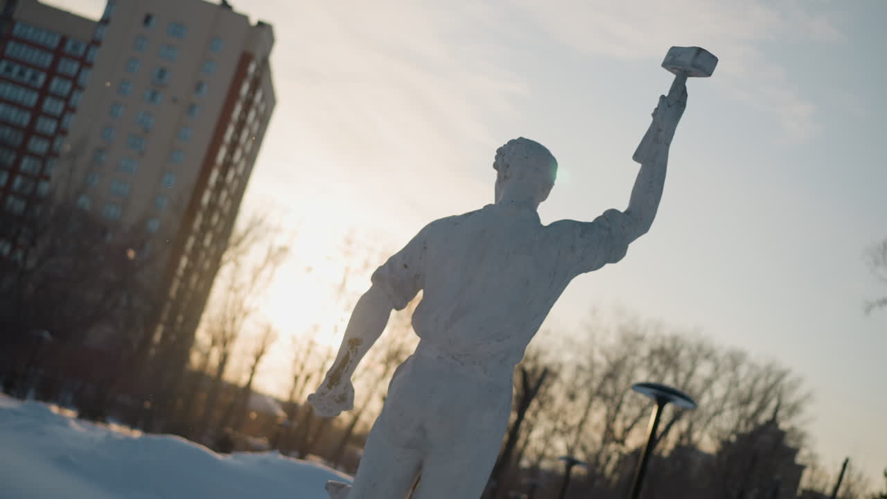 Back view of white sculpture standing in snow as sunlight creates green lens flare around statue, with high-rise buildings, bare trees, and street lamps silhouetted against cold winter sky