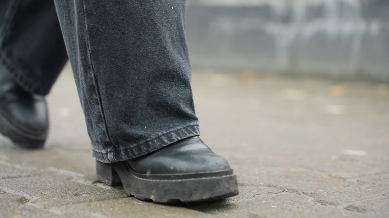 Close up of human leg wearing black trousers and black boots walking slowly forward on outdoor paved path during calm overcast day, focusing on shoe motion, wet surface, and quiet city rhythm