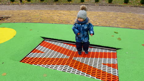 lovely toddler wearing warm jacket, cap and pants jumps on the trampoline. happy kid spending active time on the playground in autumn.