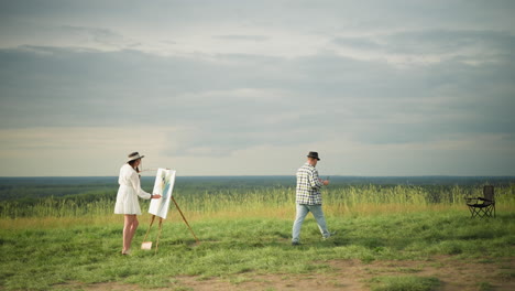 a woman in a white dress focuses on painting a canvas with a palette in hand, while an artist in a black hat and checkered shirt walks away, heading toward a chair in a grass field under a cloudy sky