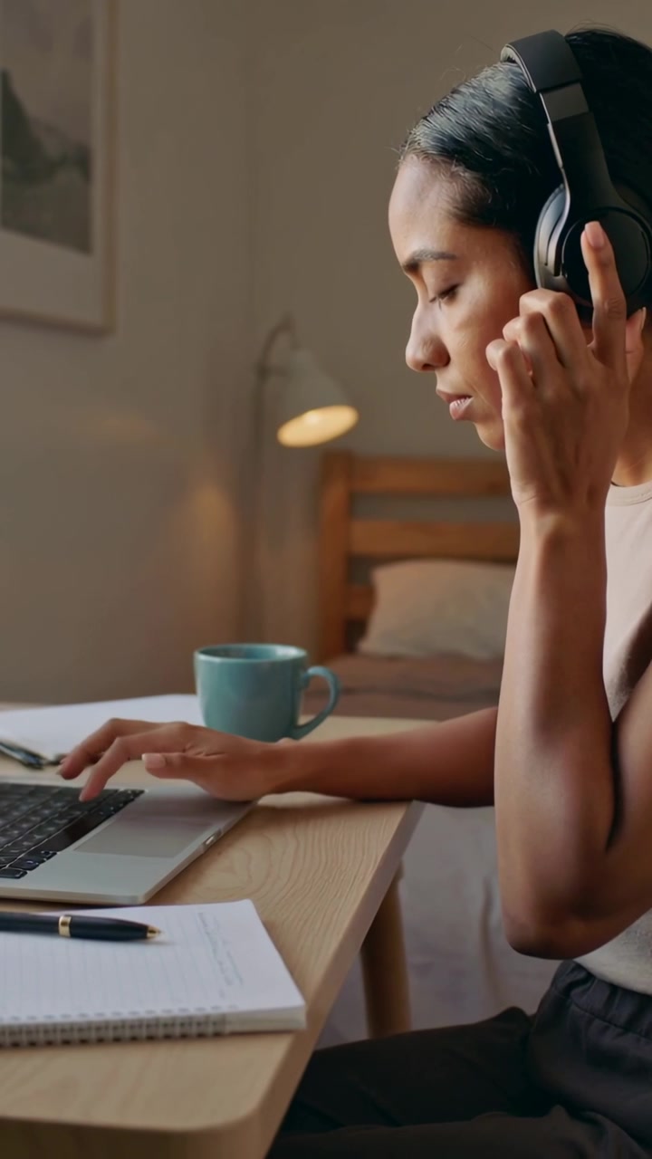 Vertical video: Adjusting headphones, woman typing on laptop at bedroom desk joining meeting