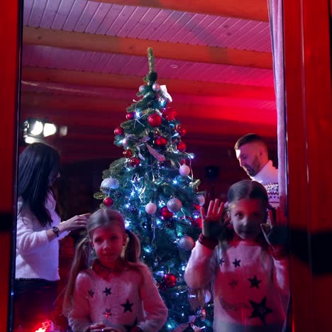 Family preparing for Christmas holidays.Parents and their children decorate New Year tree. Little girls looking through the window at Christmas eve. View from outside
