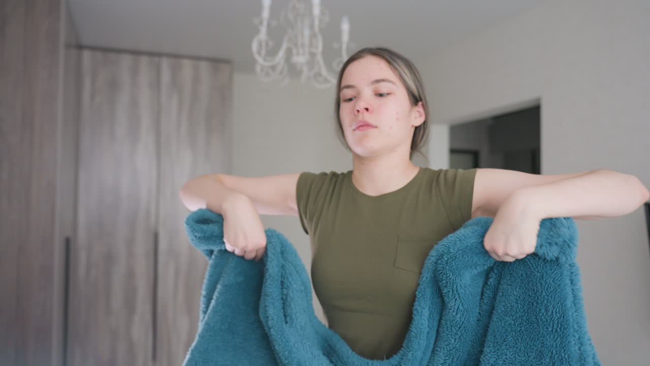 Close up of woman holding soft blue duvet while tidying living room, arranging blanket carefully with both hands, creating neat organized space illuminated by natural light through window curtains