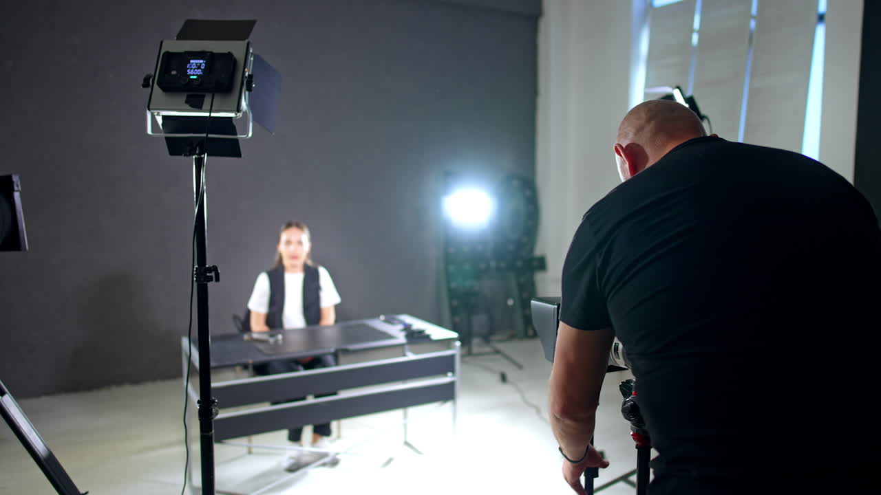 Broad-shouldered male photographer wearing black t-shirt stands his back to camera. Cameraman setting the equipment for footage of a lady sitting at desk at blurred backdrop.
