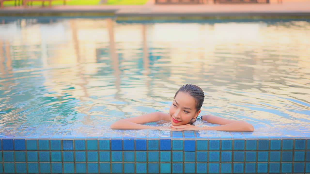 หญิงสาวเอเชียสวยเดินเข้าใกล้ขอบสระว่ายน้ำและพิงขอบสระพร้อมรอยยิ้ม แสดงถึงการพักผ่อนในโรงแรมเขตร้อน