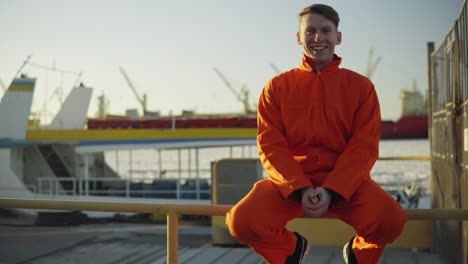 Junger-Mann-In-Orangefarbener-Uniform-Sitzt-Während-Seiner-Pause-Am-Meer-Im-Hafen-Auf-Dem-Zaun