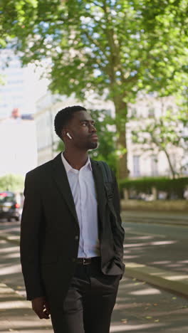 vertical video of young businessman wearing wireless earbuds streaming music or podcast from mobile phone walking to work in offices in the financial district of the city of london uk