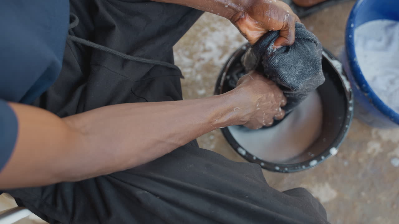 Uomo di colore che lava i panni in un secchio, primo piano di mani insaponate che strofinano un tessuto scuro sopra un catino di plastica su un portico di cemento, atmosfera di faccende domestiche mattutine, schiuma e goccioline d'acqua, tessuto con trama.