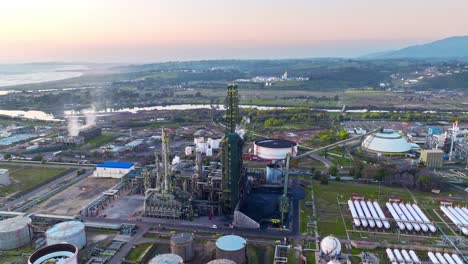 aerial orbit of a fractionation tower at an oil refinery in the middle of a natural landscape at sunset