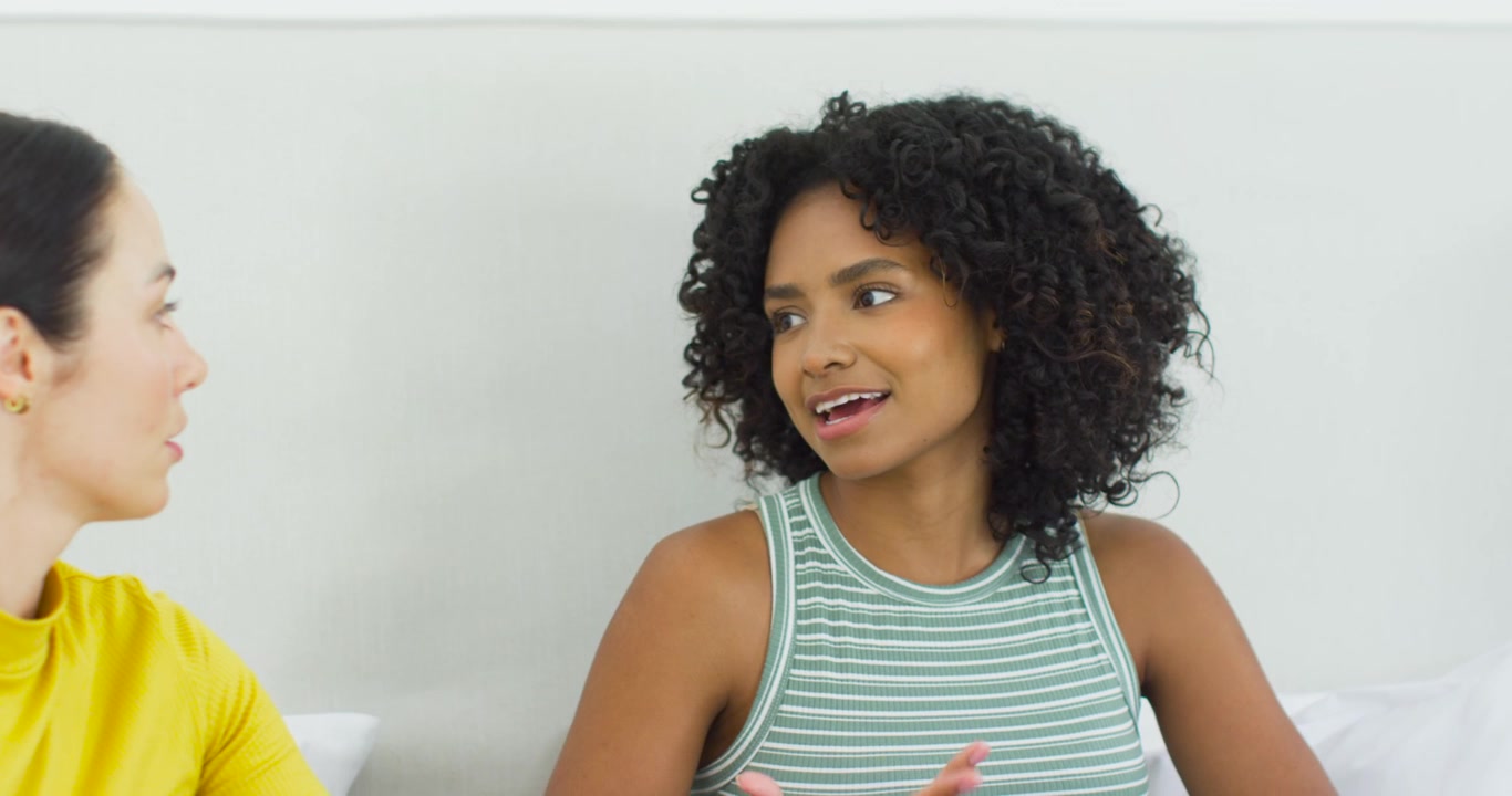 Female friends talking on couch with pillow, yellow top asking, striped woman responding, gesturing