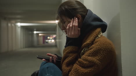 a close-up shot of a girl in a brown coat, sitting bent over in an underpass tunnel, scrolling through her phone with a heavy-hearted expression. the scene captures a moment of deep sadness