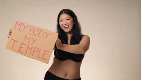 검은 속옷을 입은 미소 짓는 플러스 사이즈 여성이 스튜디오에서 춤을 추고 배너를 들고 있습니다.