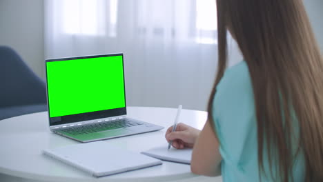 Woman-sits-at-desk-in-bedroom-she-looks-at-laptop-green-screen-and-talks-to-someone-over-internet-video-communications-sometimes-taking-notes-in-notebook.-Close-up