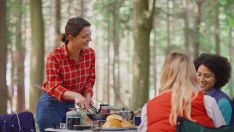 group of female friends on camping holiday in forest cooking meal sitting by tent together