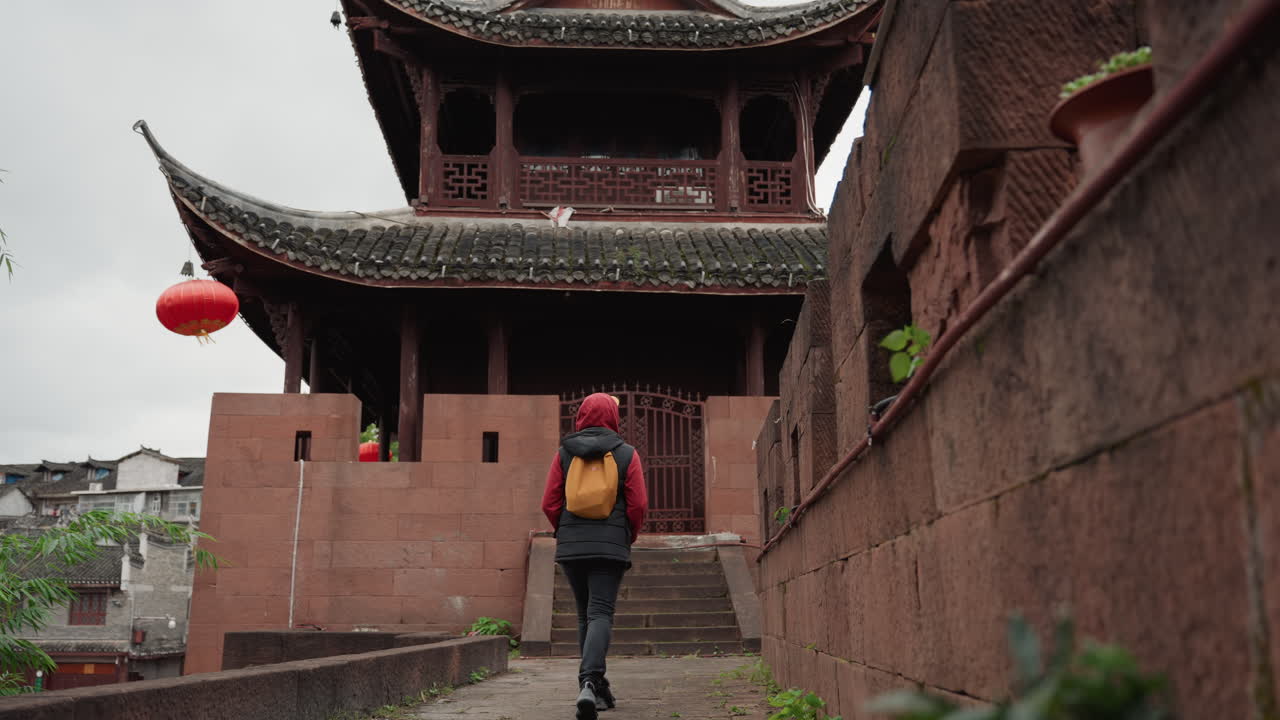 Viaggiatore in giacca rossa si avvicina al tempio, cielo coperto, scale in pietra che conducono a una pagoda ornata, lanterna rossa appesa alla grondaia, cortile stretto e muri di mattoni, backpacker che esplora un villaggio storico.