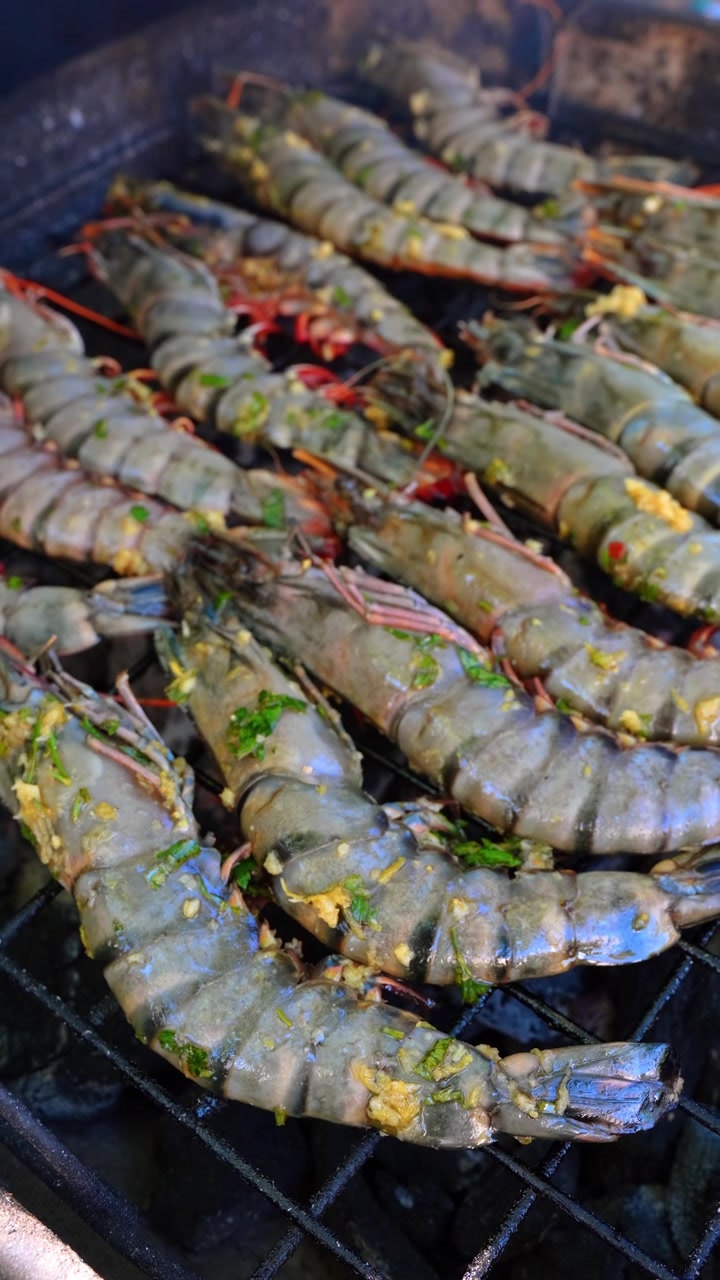Raw giant prawns cooking on outdoor barbecue grill. Gray and blue shellfish coated with chopped garlic and green herbs, viewed close up on black grates.