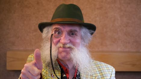 hombre mayor con barba, monóculo y sombrero, sonriendo y pulgares hacia la cámara