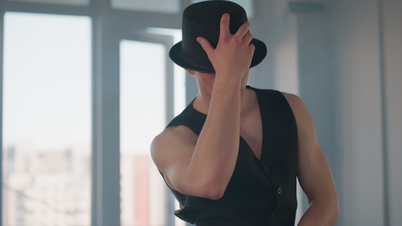 handsome man in tailored black vest and matching hat in sunlit studio removes hat brushes hair then replaces hat with confident tilt before launching into smooth dance routine against towering window