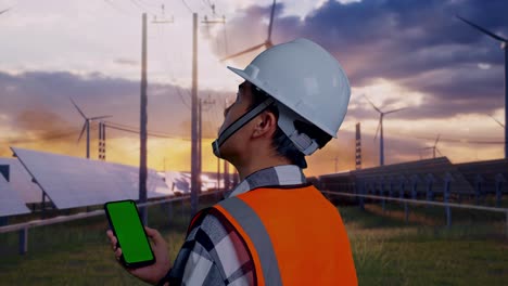 close up back view of asian male engineer with safety helmet working on a green screen smartphone and looking around while standing with solar panel and wind turbines