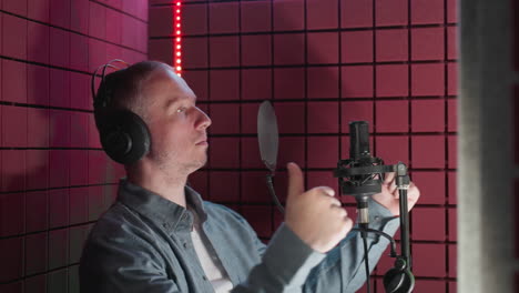 a man in a blue shirt and white inner shirt is in a red soundproof room, wearing headphones, and moving his body while singing into a microphone