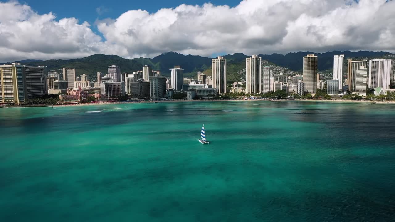 Vista aerea da drone dello skyline di Waikiki Beach con grattacieli, acqua turchese dell'oceano e barca a vela vicino alla riva sotto un cielo azzurro e nuvole bianche a Honolulu, Hawaii