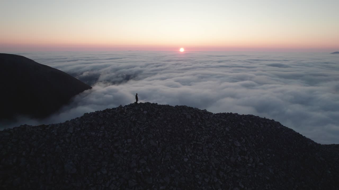 Male standing on a mountain during a colorful sunset over the valley covered in clouds. Aerial drone panning