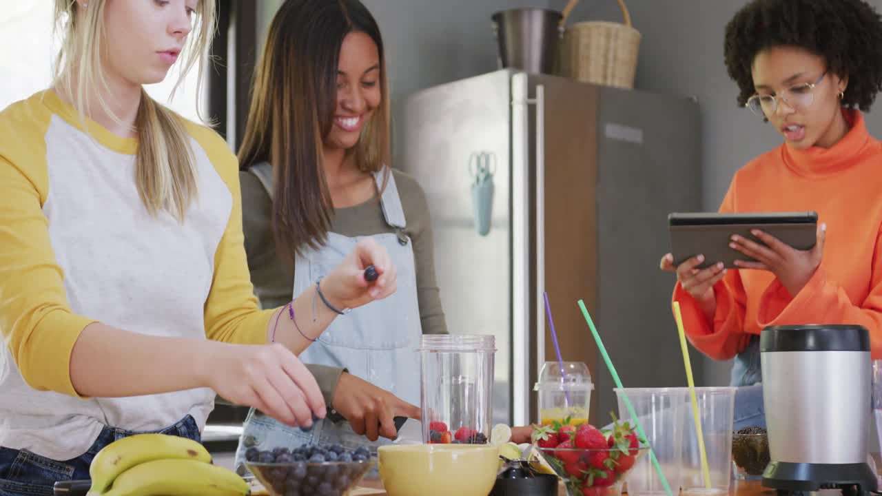 niñas adolescentes felices y diversas amigas preparando bebidas saludables usando tableta en la cocina, cámara lenta