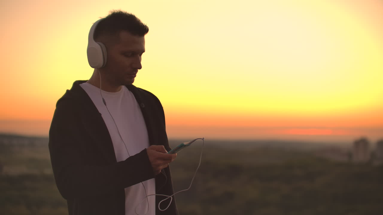 Smiling and laughing handsome young man listening music from his smartphone in wireless headphones. In slow motion a man stands on the roof at sunset and looks at the beautiful view from the height of the city