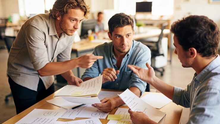 Approaching man leaning on desk pointing and sliding documents for review in office stickies shirts