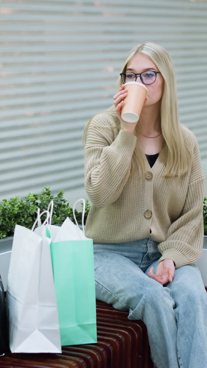 뜨개질 스웨터를 입고 컵에서 커피를 마시는 긴 머리카락의 여인이 쇼핑몰에 앉아 있으며, 배경에는 세련된 의류와 액세서리를 선보이는 옷 가게가 있습니다.