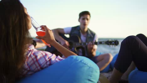 group of friends drinking cocktails and beer and doing cheers sitting on easychairs on the beach and listening to a friend playing guitar on a summer evening during a sunset. slowmotion shot