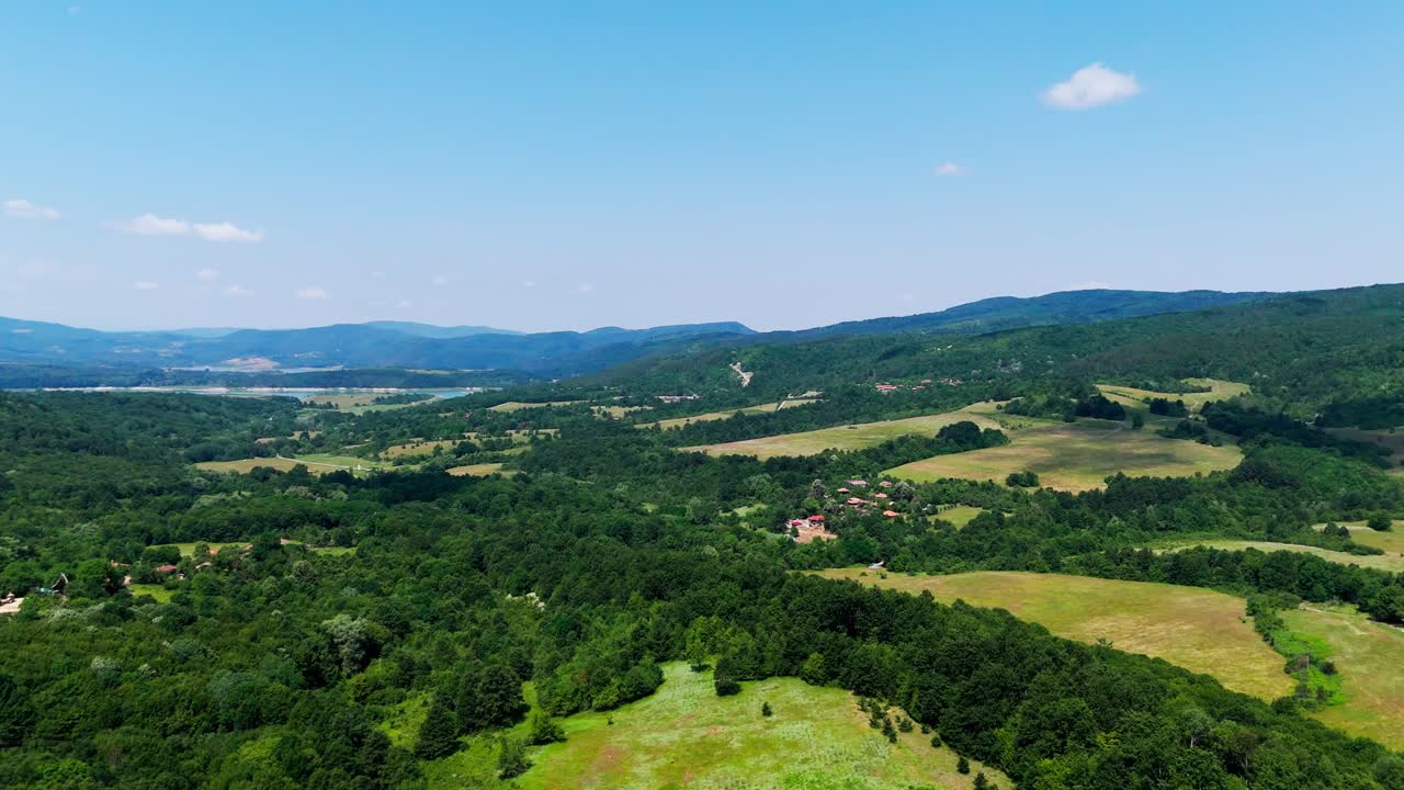 Ampia panoramica aerea che rivela colline balcaniche, vegetazione, case, villaggi, un lago e montagne lontane e sfocate, sotto un cielo azzurro.